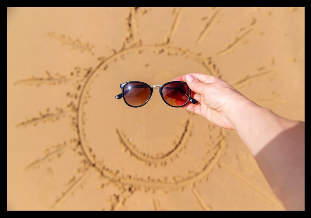 V písku nakreslené sluníčko, ruka držící brýle v místě kde má sluníčko oči